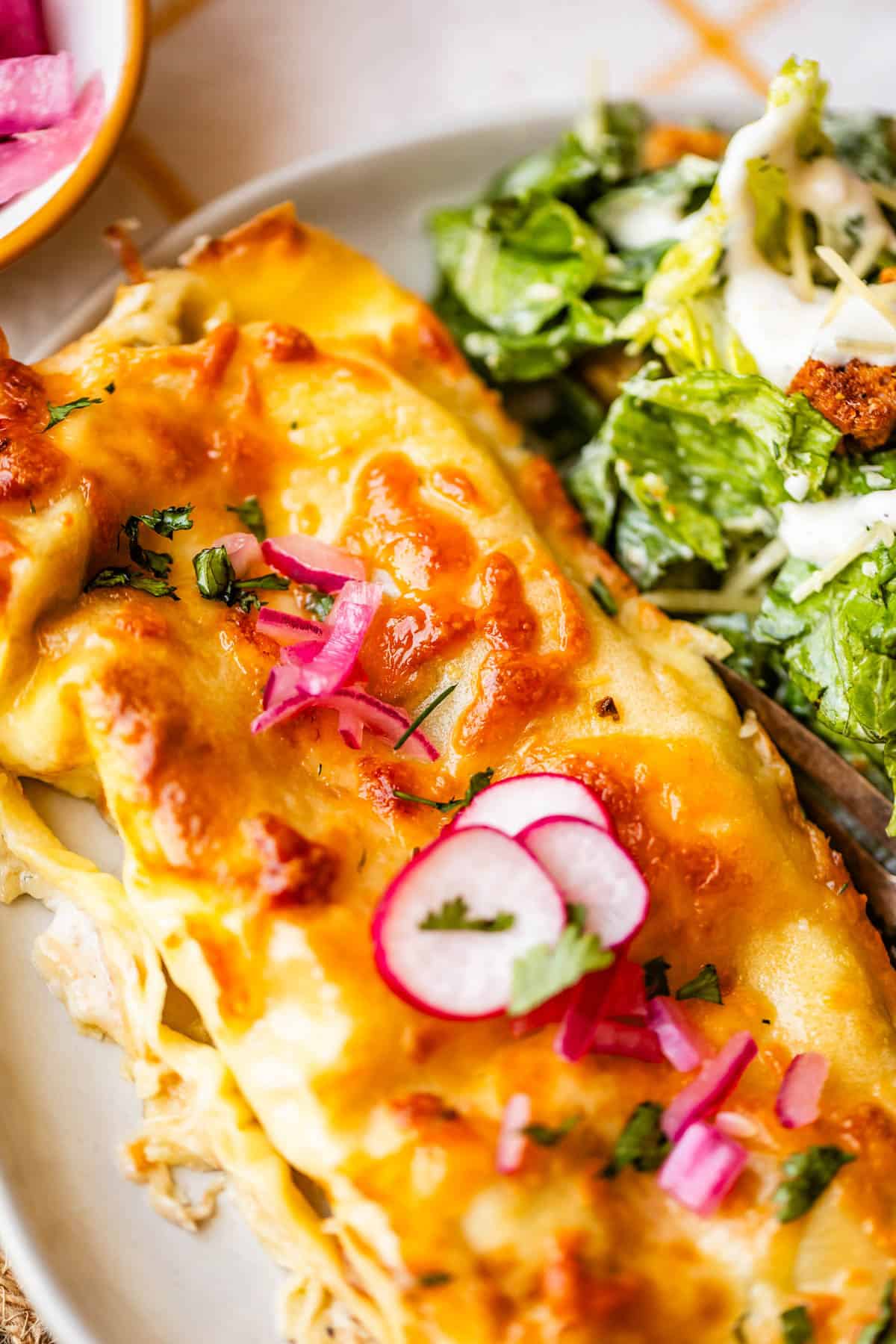 A close-up of cheesy enchiladas topped with radish slices, pickled onions, and cilantro, served next to a green salad with creamy dressing.