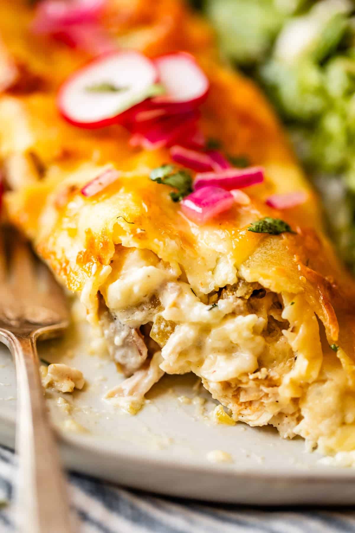 A close-up of a cheesy chicken enchilada topped with sliced radishes and pickled onions, with a fork beside it and a bite taken out, revealing the creamy filling inside.