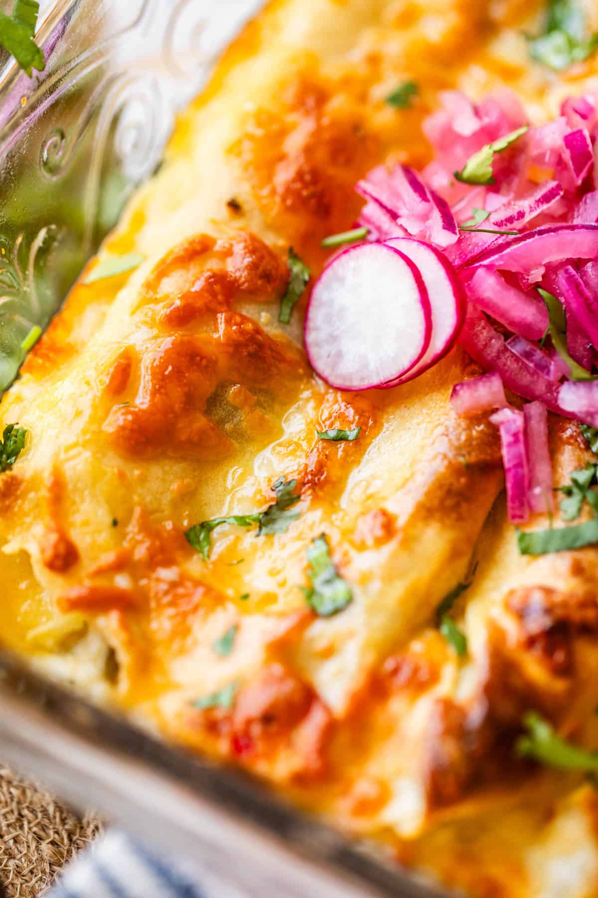 Close-up of cheesy baked enchiladas topped with fresh chopped cilantro, thinly sliced radishes, and pickled red onions in a glass baking dish.