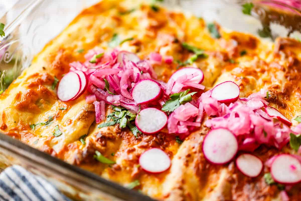 Close-up of baked enchiladas in a glass dish, topped with melted cheese, sliced radishes, pickled red onions, and fresh cilantro.