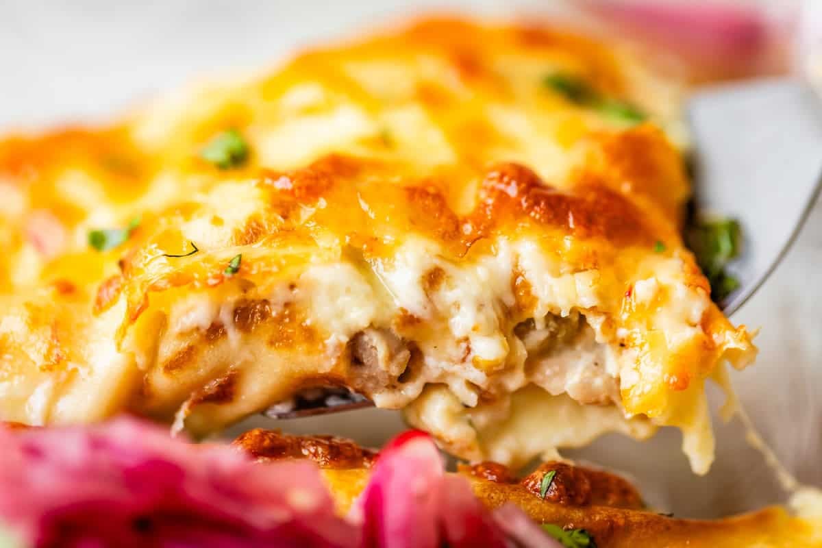 Close-up of a cheesy, golden-brown slice of lasagna with visible layers of meat, sauce, and melted cheese, being served with a spatula. Pink pickled onions are visible in the foreground.