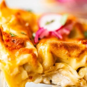 Close-up of a cheesy lasagna slice with golden-brown crust, creamy layers, and topped with sliced red onions and herbs. The lasagna is being lifted from a glass baking dish.