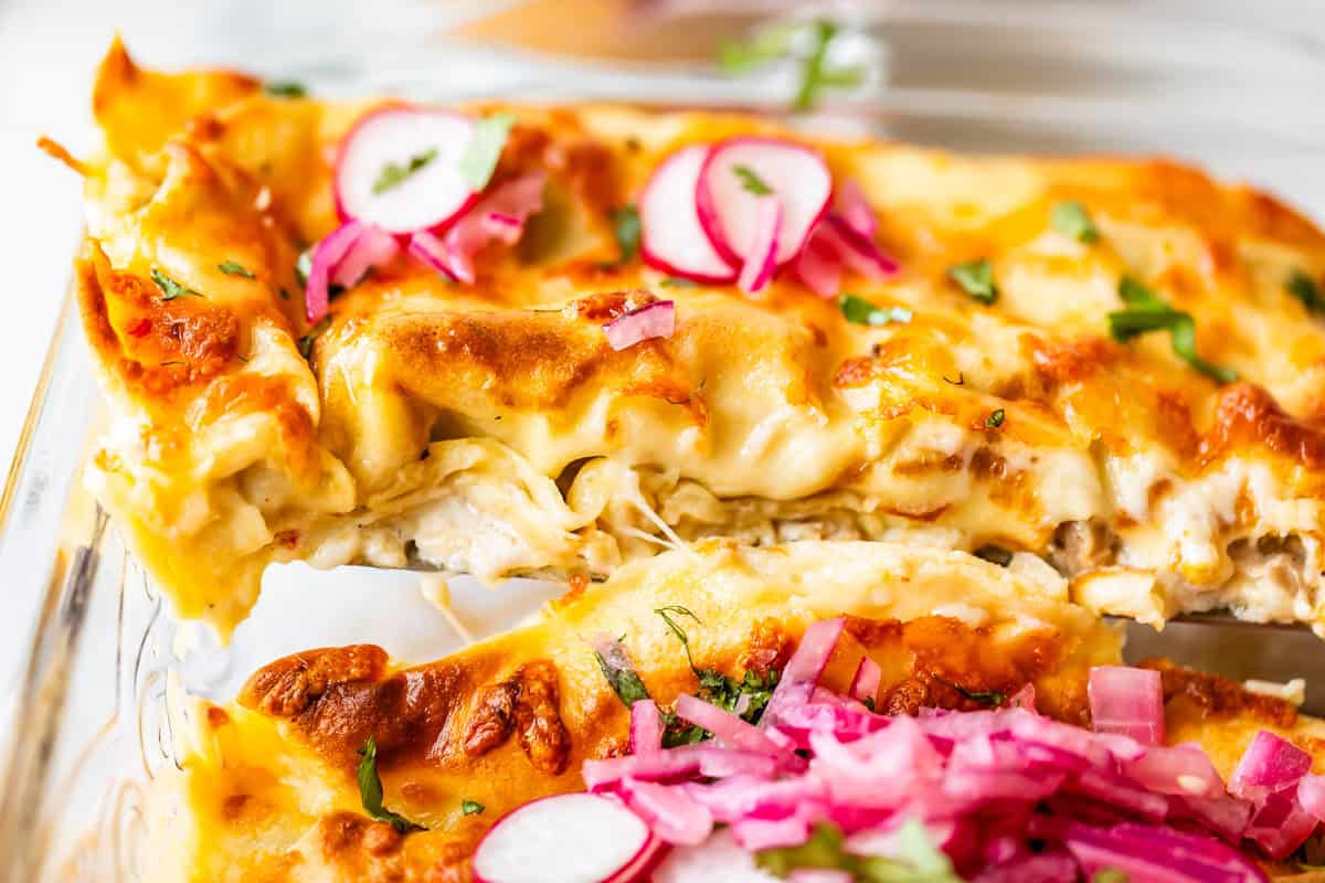 A close-up of cheesy baked lasagna topped with sliced radishes and pickled onions in a glass dish, showing melted golden cheese and herbs.