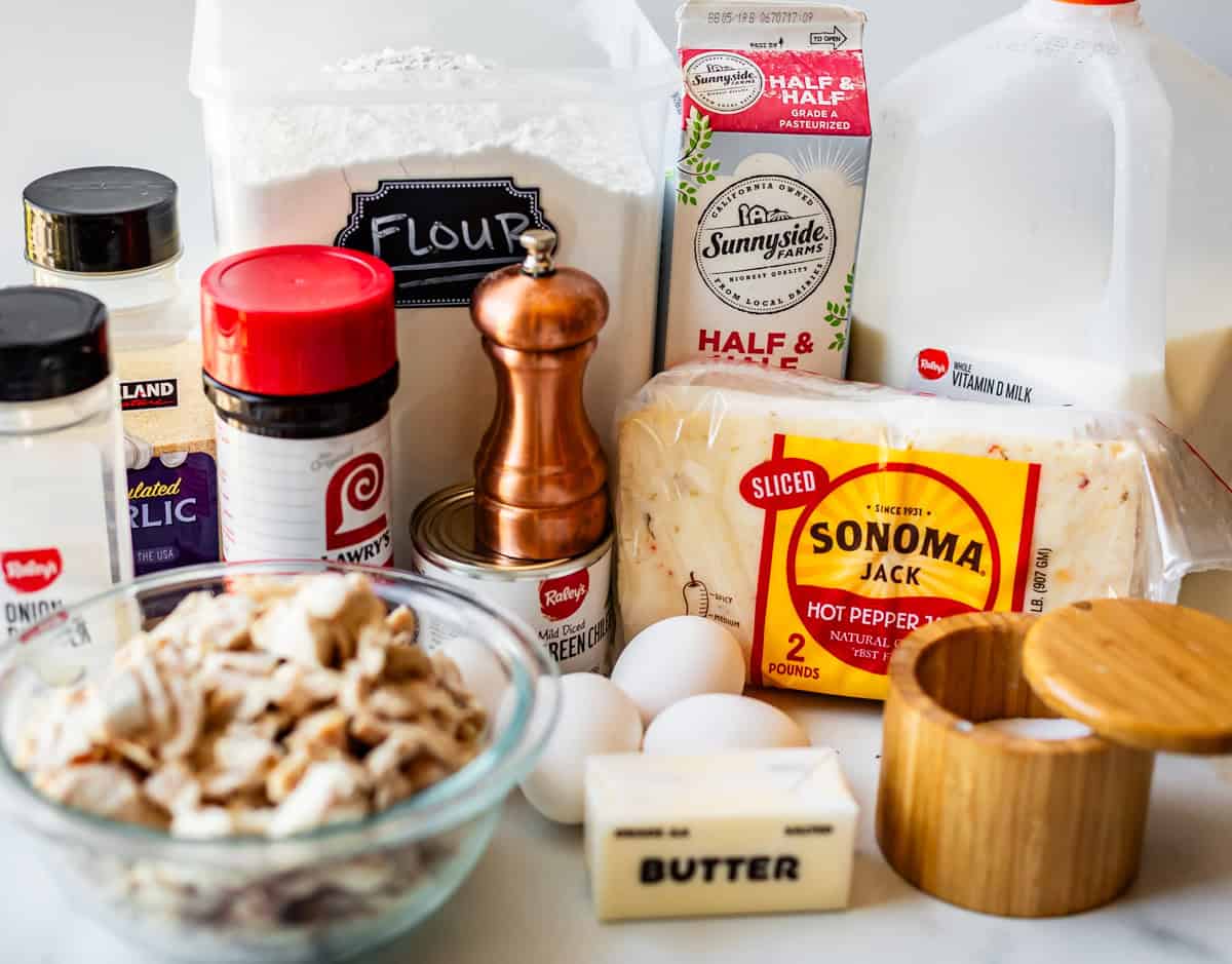 A variety of cooking ingredients on a counter, including flour, half & half, milk, sliced hot pepper jack cheese, eggs, butter, seasonings, pepper grinder, a bowl of chopped chicken, and containers of garlic and onion powder.