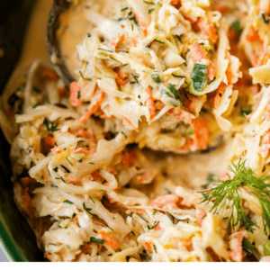 Close-up of creamy homemade coleslaw with shredded cabbage, carrots, and dill in a bowl, with a spoon for serving. Text reads: The Best Homemade Easy Coleslaw. The Food Charlatan.