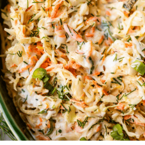 A close-up of creamy coleslaw with shredded cabbage, carrots, green onions, and herbs in a green bowl. Text at the bottom reads “The Best Homemade Easy Coleslaw The Food Charlatan.”.