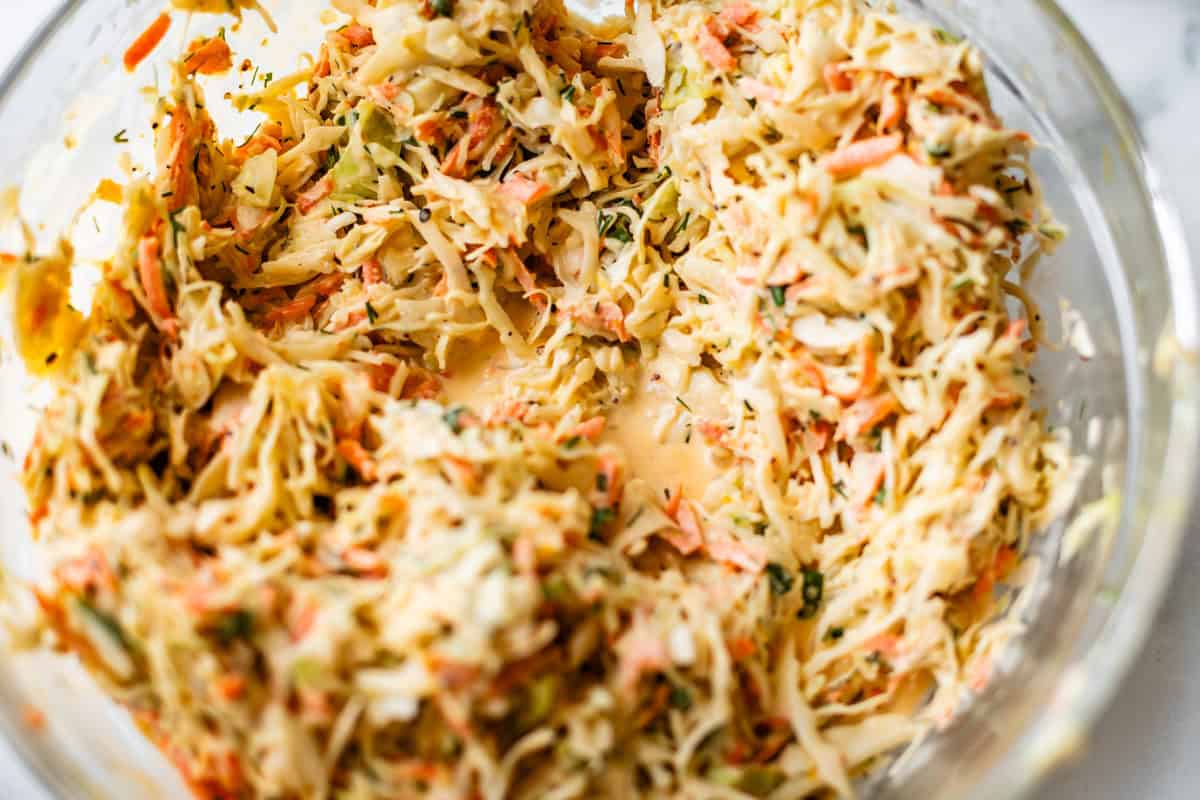A close-up of a glass bowl filled with creamy coleslaw, featuring shredded cabbage, carrots, and herbs mixed with a dressing.