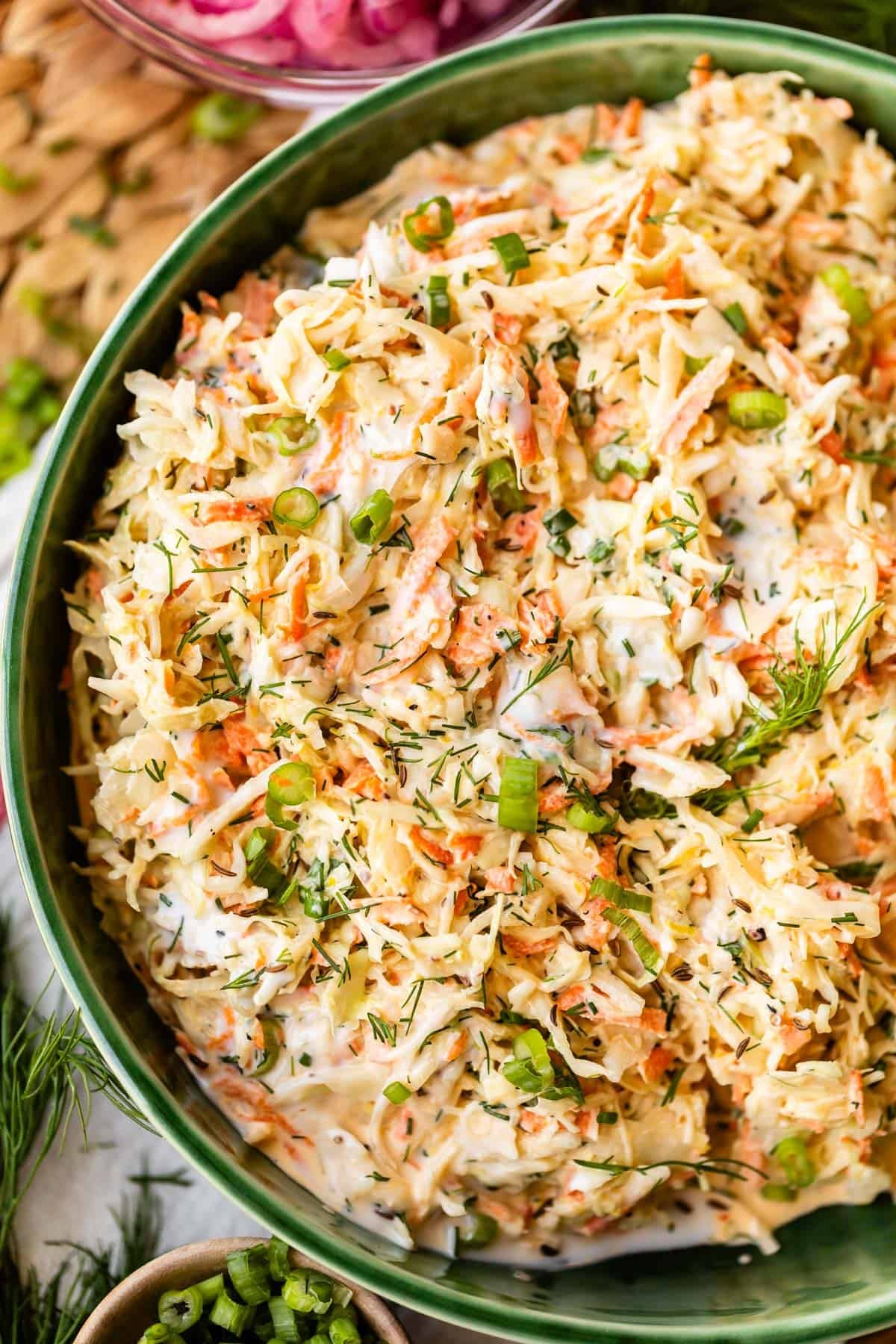 A close-up of a bowl filled with creamy coleslaw, featuring shredded cabbage, carrots, and green onions, garnished with fresh dill.