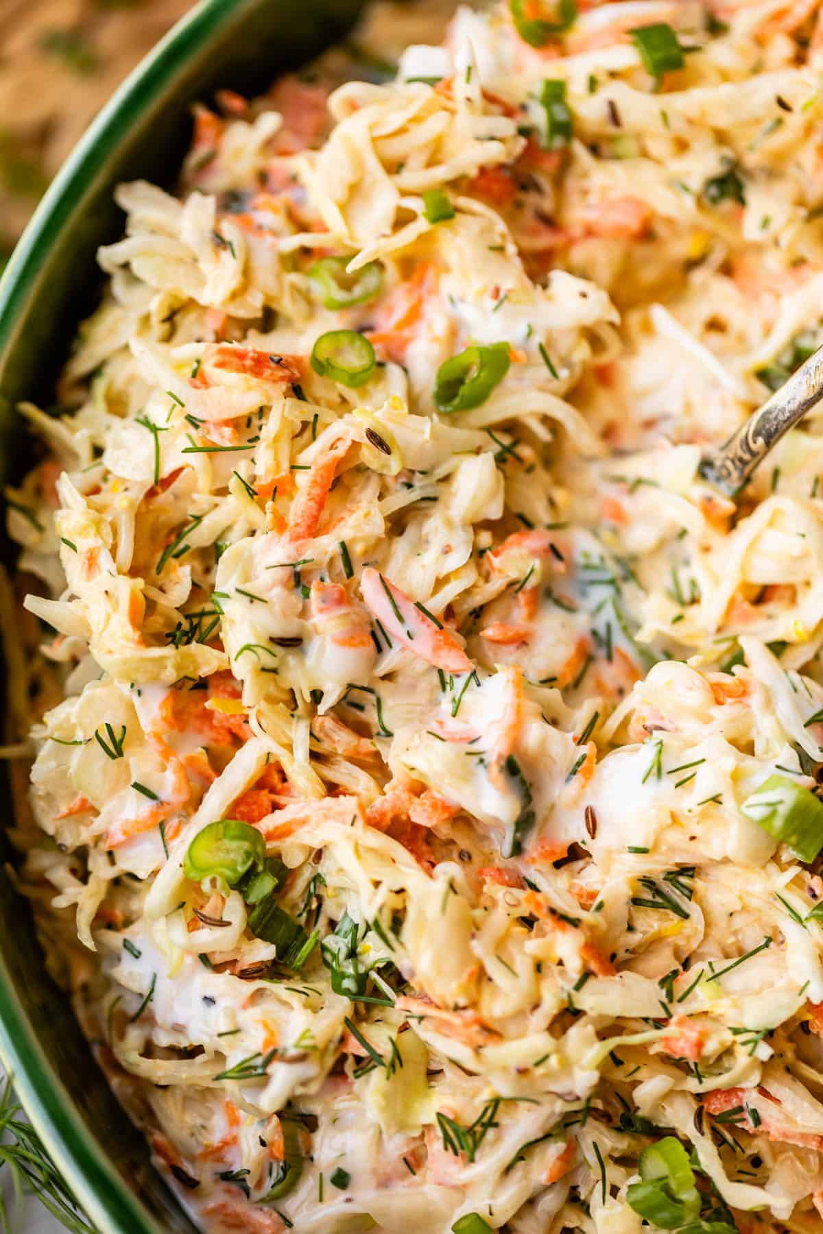 Close-up of creamy coleslaw with shredded cabbage, carrots, chopped green onions, and fresh herbs, all mixed together in a bowl with a silver spoon.
