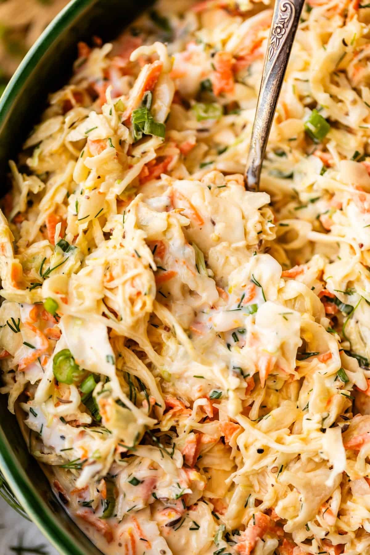 A close-up of creamy coleslaw with shredded cabbage, carrots, green onions, and fresh herbs in a bowl with a serving spoon.