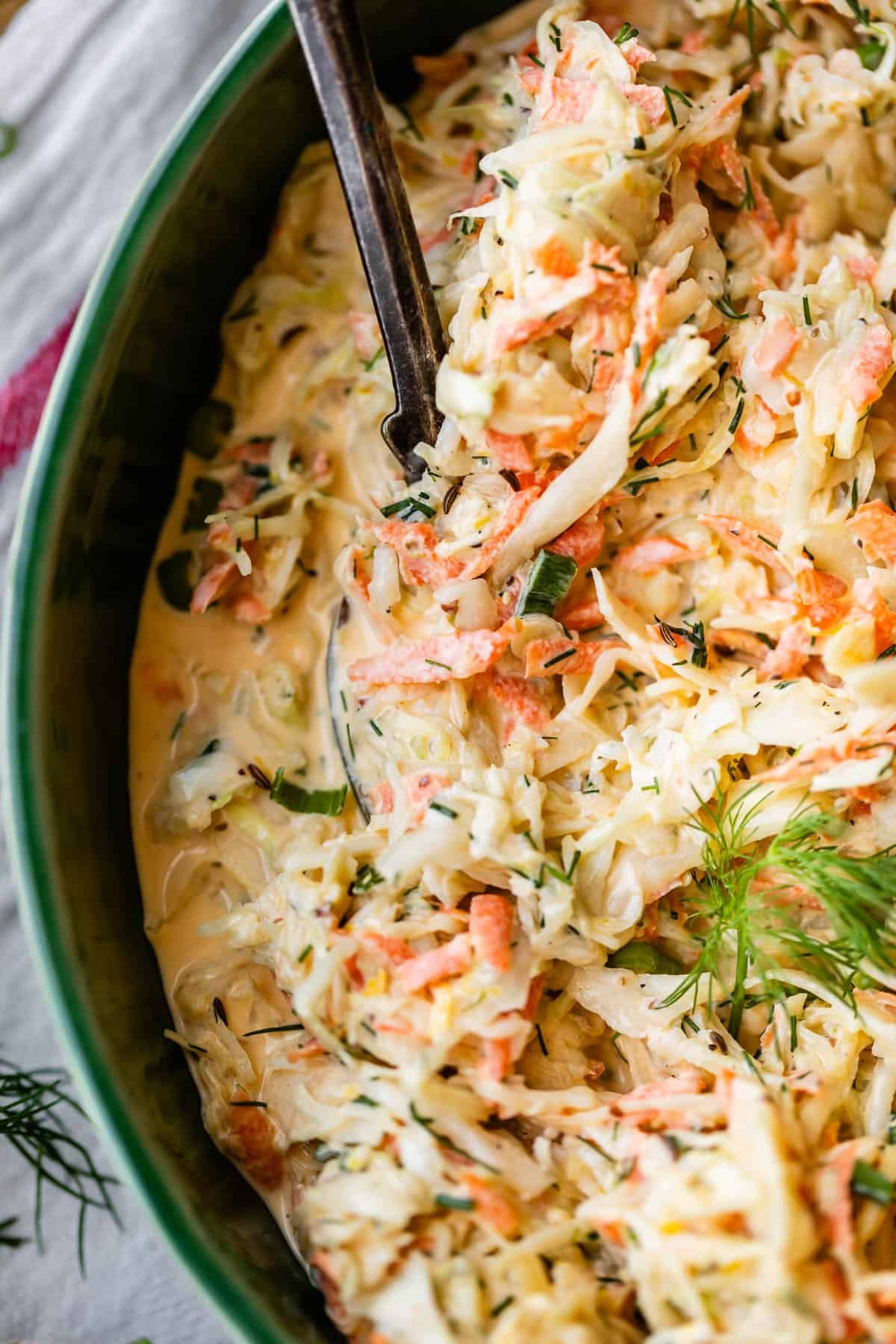 Close-up of creamy coleslaw with shredded cabbage, carrots, fresh dill, and a mayonnaise-based dressing in a green bowl, with a serving spoon partially visible.