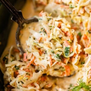 A close-up of creamy coleslaw being served with a spoon, showing shredded cabbage, carrots, herbs, and a creamy dressing. A sprig of dill garnishes the dish.