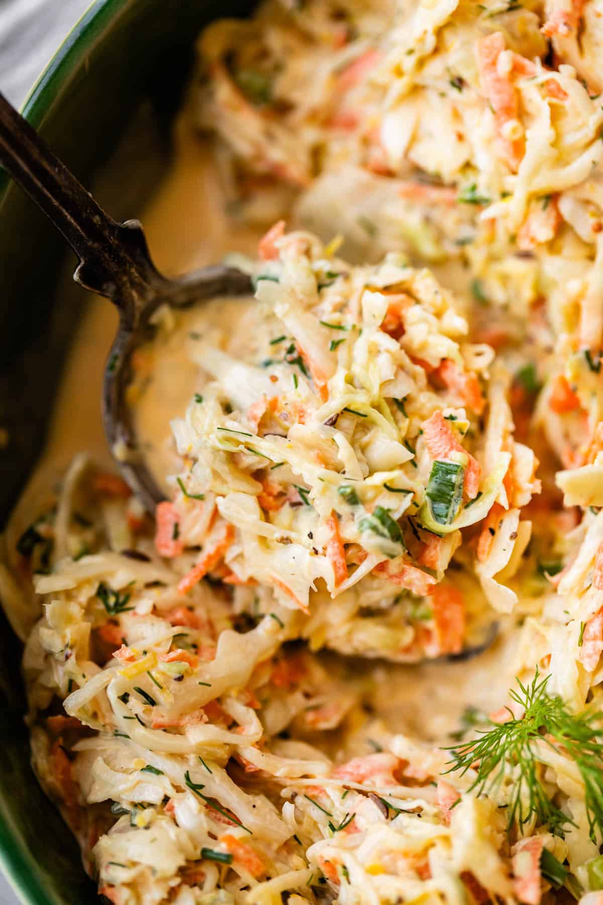 A close-up of creamy coleslaw being served with a spoon, showing shredded cabbage, carrots, herbs, and a creamy dressing. A sprig of dill garnishes the dish.