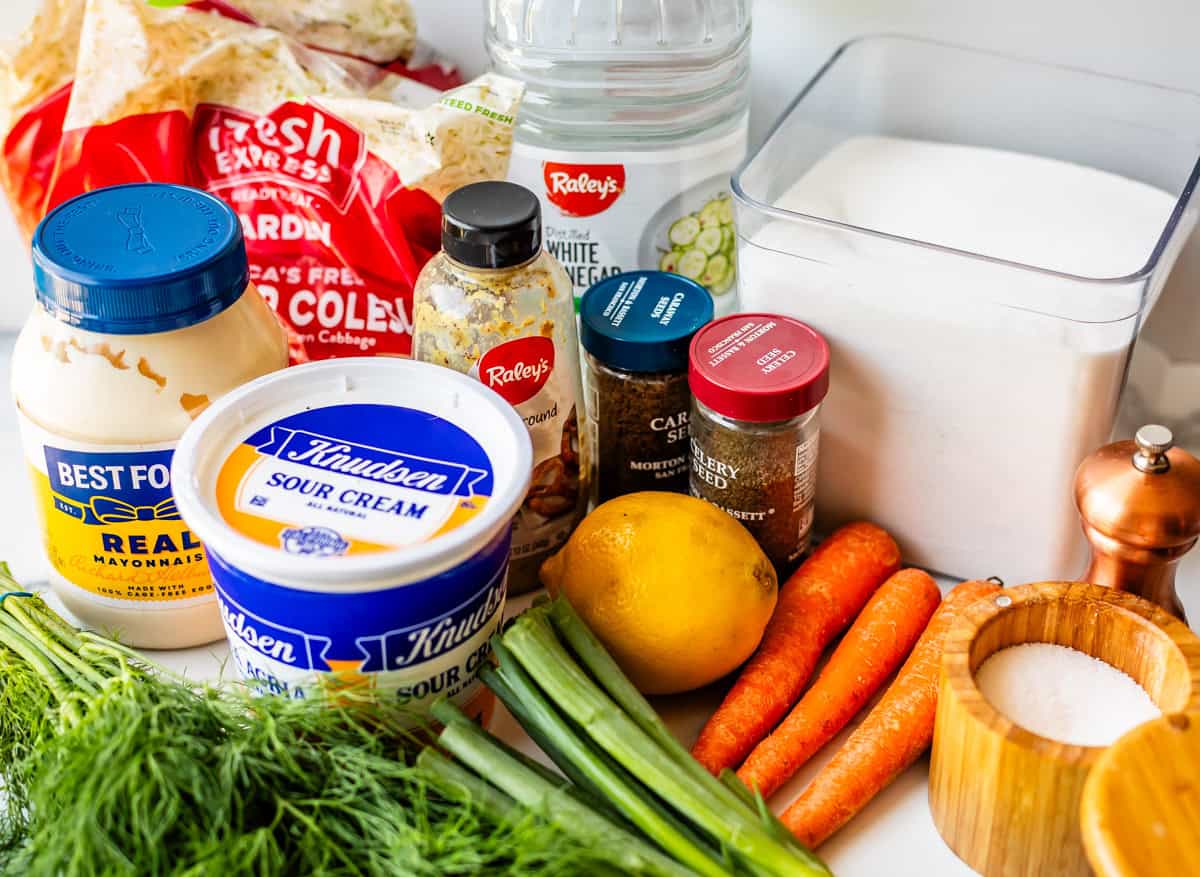 An assortment of coleslaw ingredients on a counter, including mayonnaise, sour cream, shredded coleslaw mix, carrots, a lemon, fresh dill, green onions, sugar, white vinegar, and various spices.