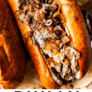 A close-up of a Philly cheesesteak sandwich filled with grilled beef, melted cheese, and caramelized onions in a toasted roll, surrounded by potato chips. Text at the bottom reads: Philly Cheese Steaks - The Food Charlatan.