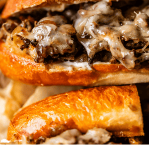 Close-up of a Philly cheesesteak sandwich with melted cheese and thinly sliced beef in a toasted roll. Text overlay reads Easy Weeknight Philly Cheesesteaks, The Food Charlatan.