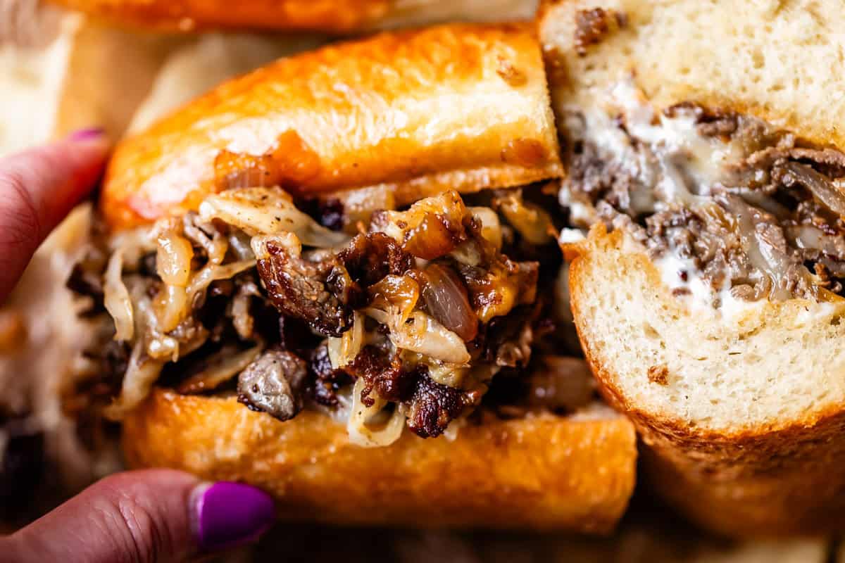 A close-up of a hand holding a cheesesteak sandwich filled with grilled steak, caramelized onions, and melted cheese, served in a toasted hoagie roll.
