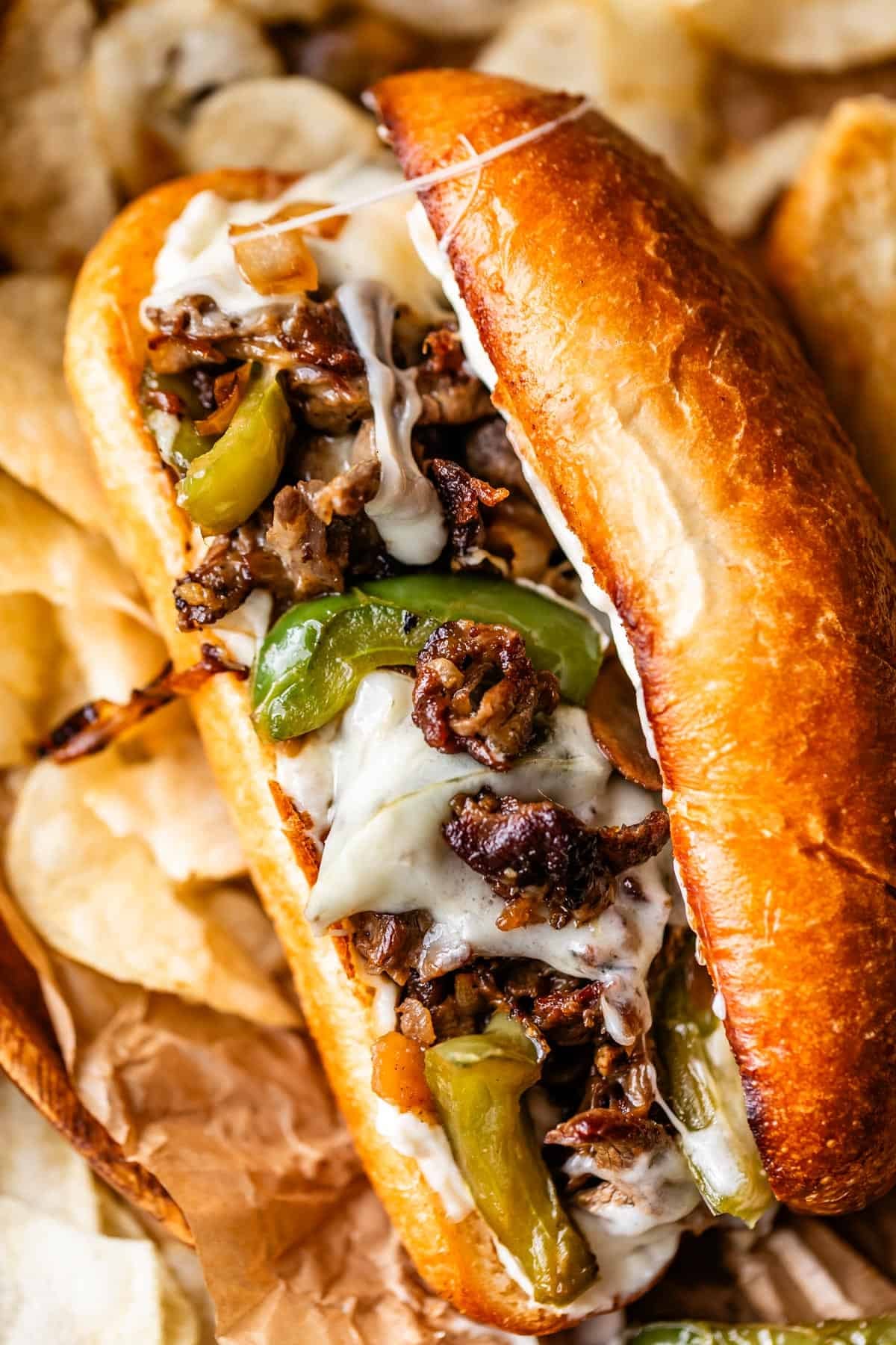 A close-up of a Philly cheesesteak sandwich on a toasted bun, filled with melted cheese, saut&eacute;ed green bell peppers, onions, and thinly sliced steak, served with potato chips in the background.