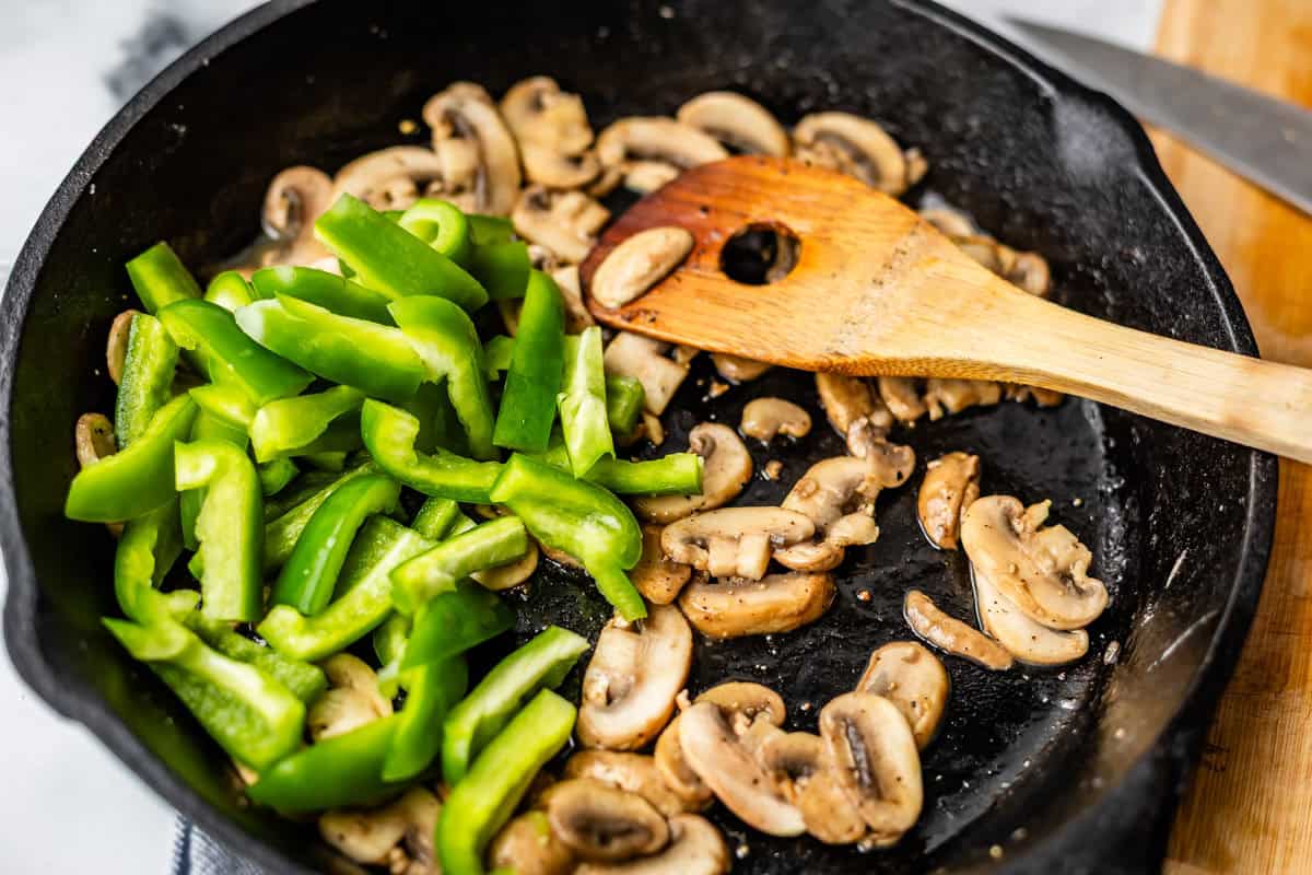 Sliced green bell peppers and mushrooms being saut&eacute;ed in a black skillet, with a wooden spatula resting inside the pan.