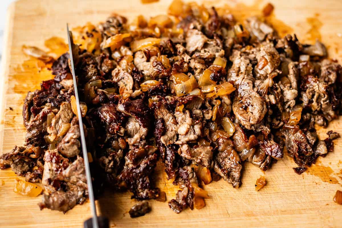 Chopped cooked beef mixed with saut&eacute;ed onions on a wooden cutting board, with a knife partially visible on the left side.