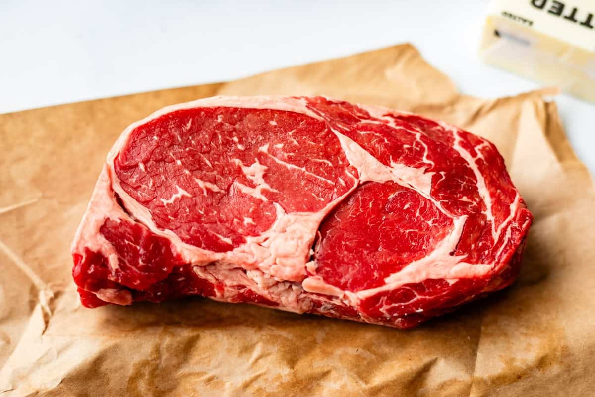 A raw ribeye steak with marbling of fat sits on a piece of brown butcher paper, with part of a stick of butter visible in the background.