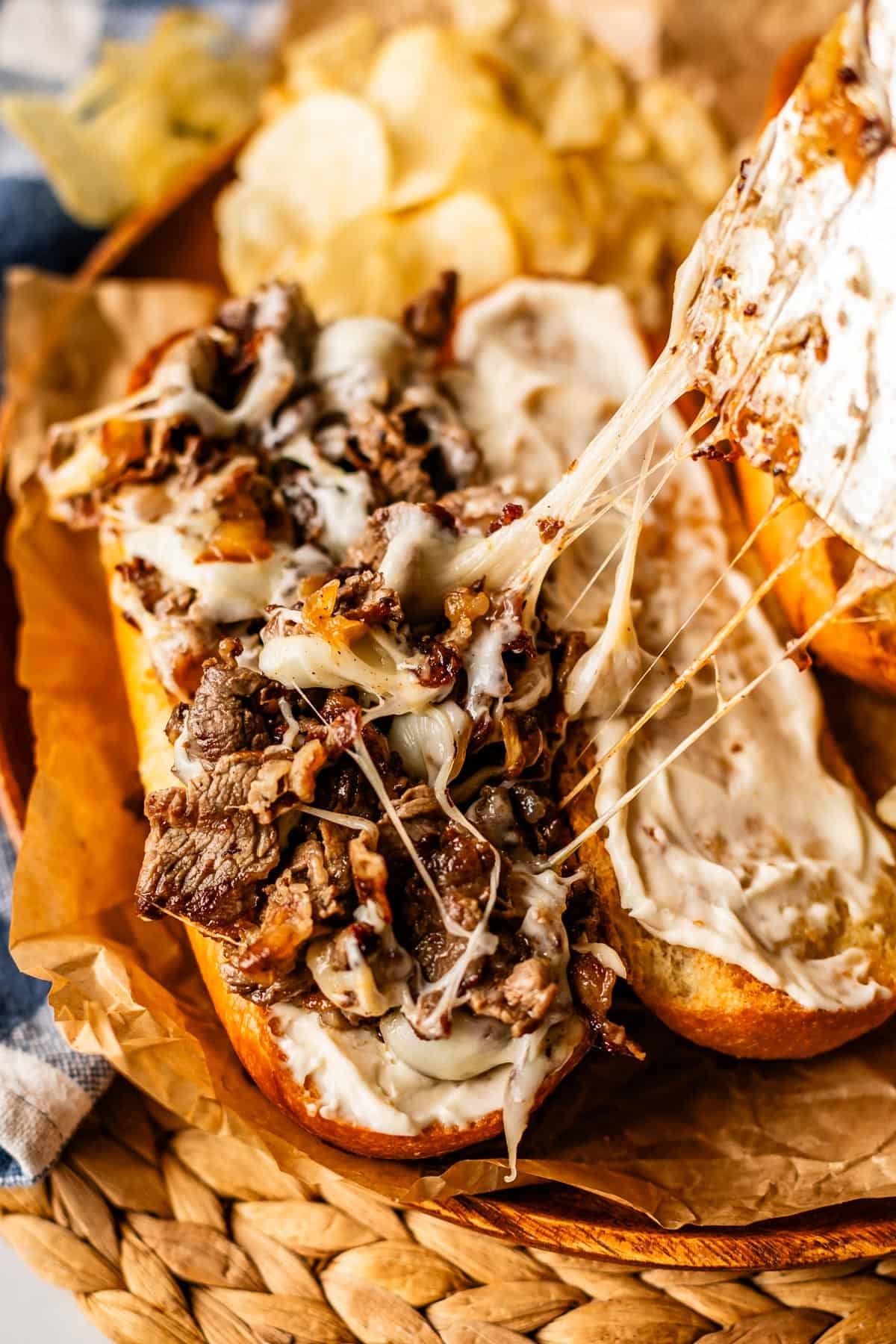 A close-up of a cheesesteak sandwich on a toasted bun, topped with melted cheese, saut&eacute;ed onions, and sliced steak, with potato chips in the background. The melted cheese is stretching as the sandwich is pulled apart.