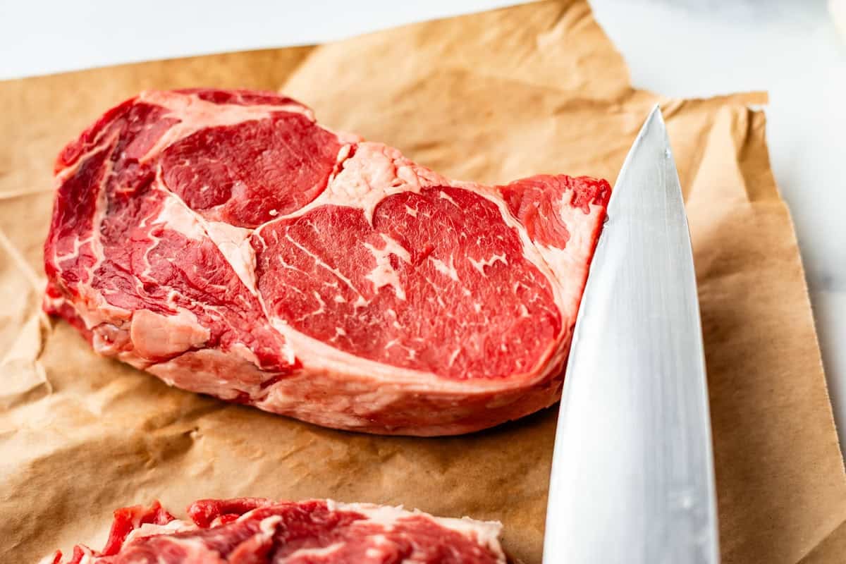A raw ribeye steak on brown parchment paper next to a large kitchen knife, ready for preparation.