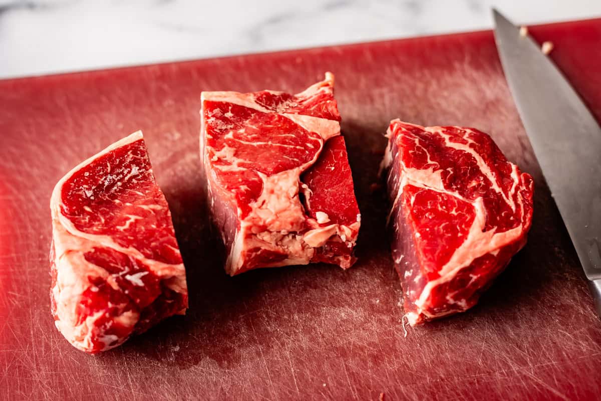 Three thick, raw beef chunks sit on a red cutting board next to a large kitchen knife; marbling and fat are visible on the meat, and the background is slightly blurred.