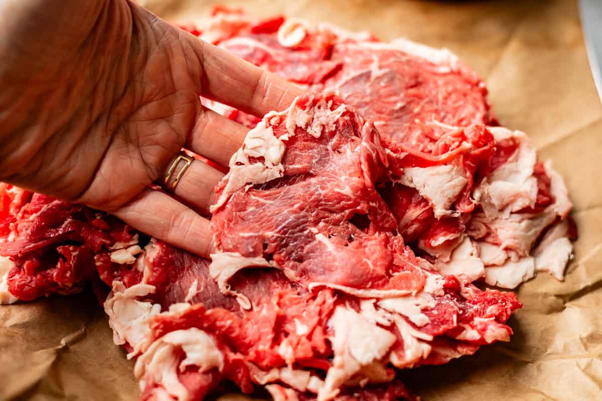 A hand holds thinly sliced raw beef with visible marbling, resting on brown parchment paper.