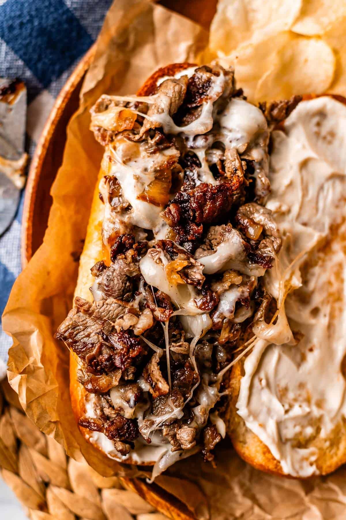 Close-up of a toasted sandwich filled with melted cheese and chopped, grilled beef, served on brown parchment paper with potato chips on the side. One half of the bread is spread with a creamy white sauce.