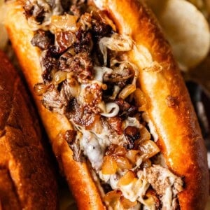 A close-up of a Philly cheesesteak sandwich filled with melted cheese, sautéed onions, and thinly sliced beef, served in a toasted bun with potato chips in the background.
