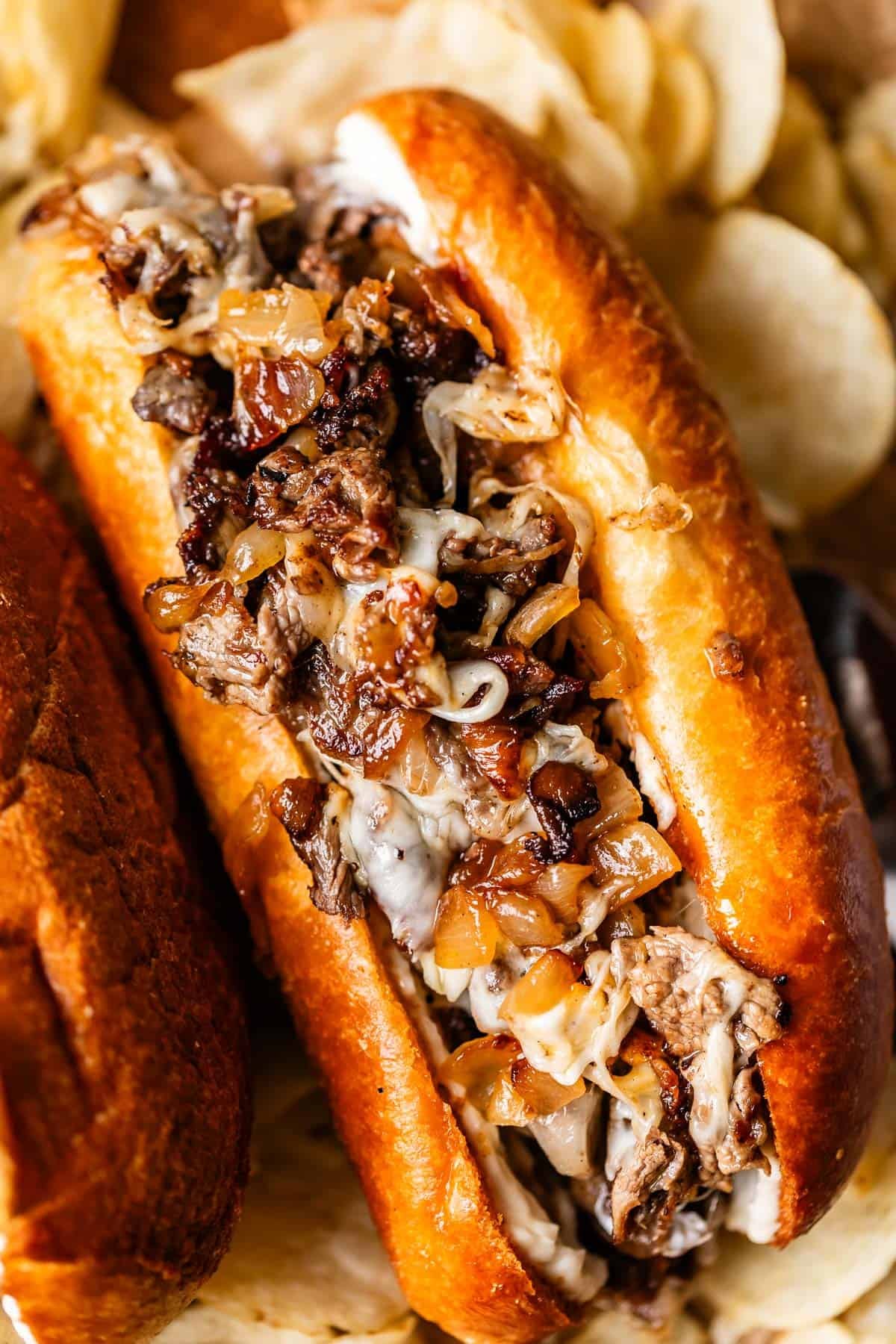 A close-up of a Philly cheesesteak sandwich filled with melted cheese, saut&eacute;ed onions, and thinly sliced beef, served in a toasted bun with potato chips in the background.