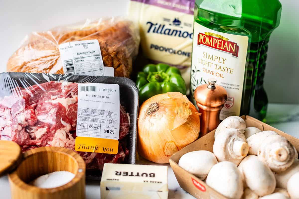 A variety of fresh ingredients on a counter, including sliced bread, provolone cheese, olive oil, beef, green bell pepper, an onion, mushrooms, butter, salt, and a pepper grinder.