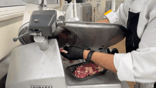 A person wearing black gloves and a white coat slices raw meat using an industrial meat slicer, collecting the slices in a tray below the machine.