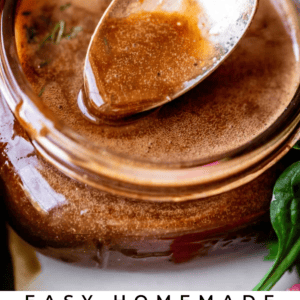A spoon rests on the rim of a jar filled with creamy balsamic vinaigrette. Fresh greens and tomatoes are visible in the background. Text below reads: Easy Homemade Creamy Balsamic Vinaigrette – The Food Charlatan.