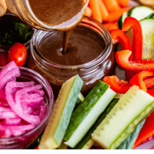 A hand pours creamy balsamic vinaigrette from a jar into a small bowl, surrounded by fresh vegetables like carrots, red peppers, cucumbers, and pickled onions. Text reads: Creamy Balsamic Vinaigrette. The Food Charlatan.
