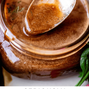 A close-up of a jar filled with balsamic vinaigrette, with a spoon resting on top. Fresh greens surround the jar. Bold text below reads, Seriously the best ever balsamic vinaigrette. The Food Charlatan.