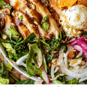 A vibrant salad with leafy greens, orange slices, grilled chicken, goat cheese, red onions, and seeds, garnished with fresh herbs.