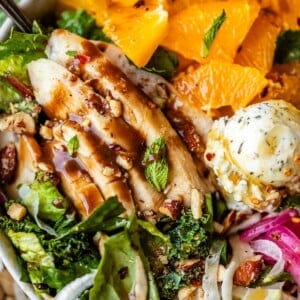 A salad bowl with sliced grilled chicken, mixed greens, orange segments, red onions, nuts, seeds, and a scoop of herb butter, all drizzled with dressing.
