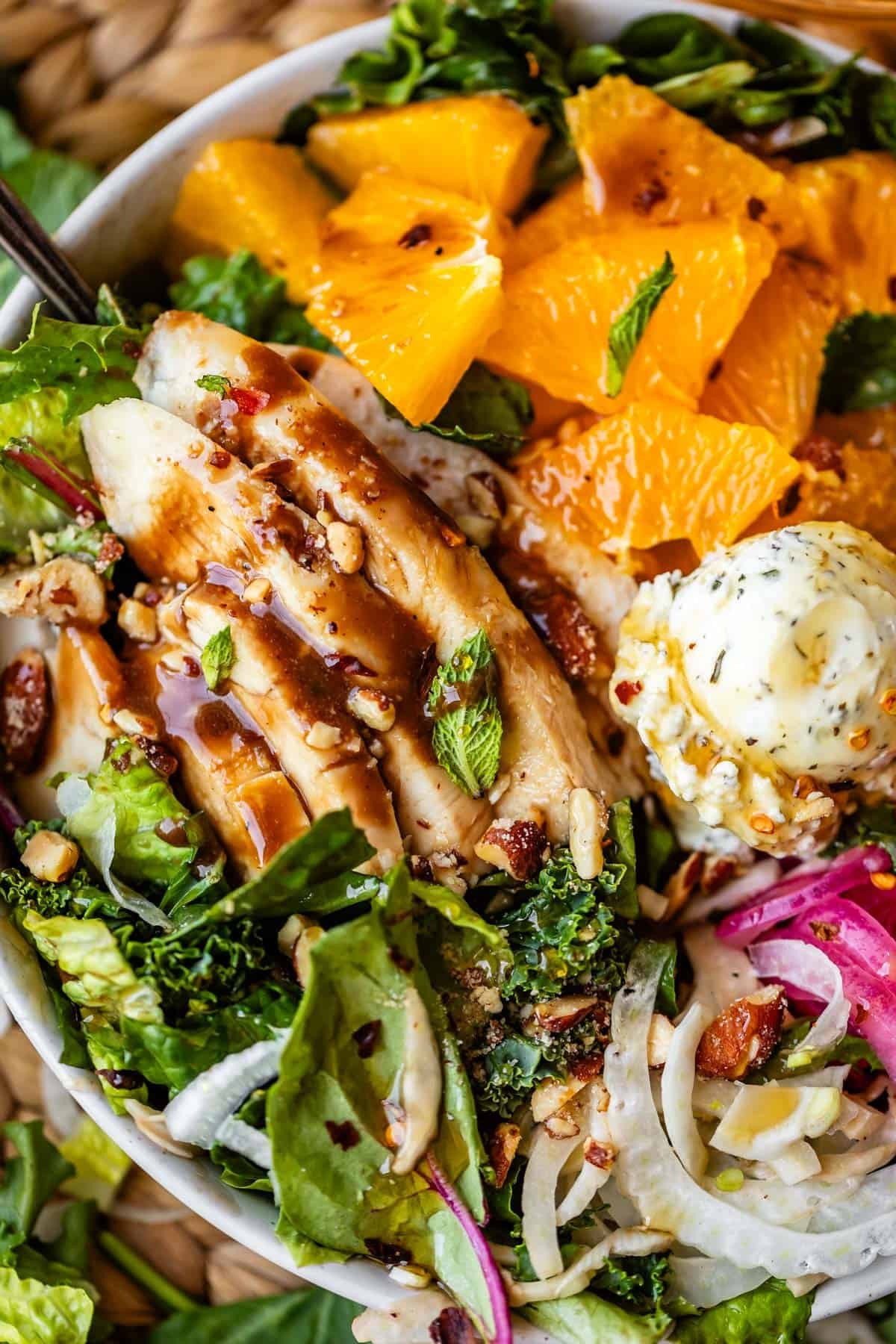 A salad bowl with sliced grilled chicken, mixed greens, orange segments, red onions, nuts, seeds, and a scoop of herb butter, all drizzled with dressing.