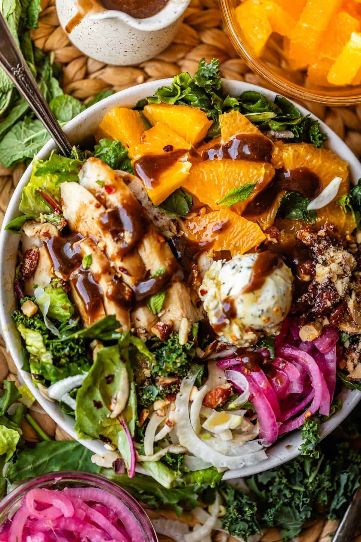 A vibrant salad bowl with orange slices, grilled chicken, pickled onions, nuts, greens, goat cheese, and dark dressing, surrounded by extra pickled onions and citrus in small bowls.