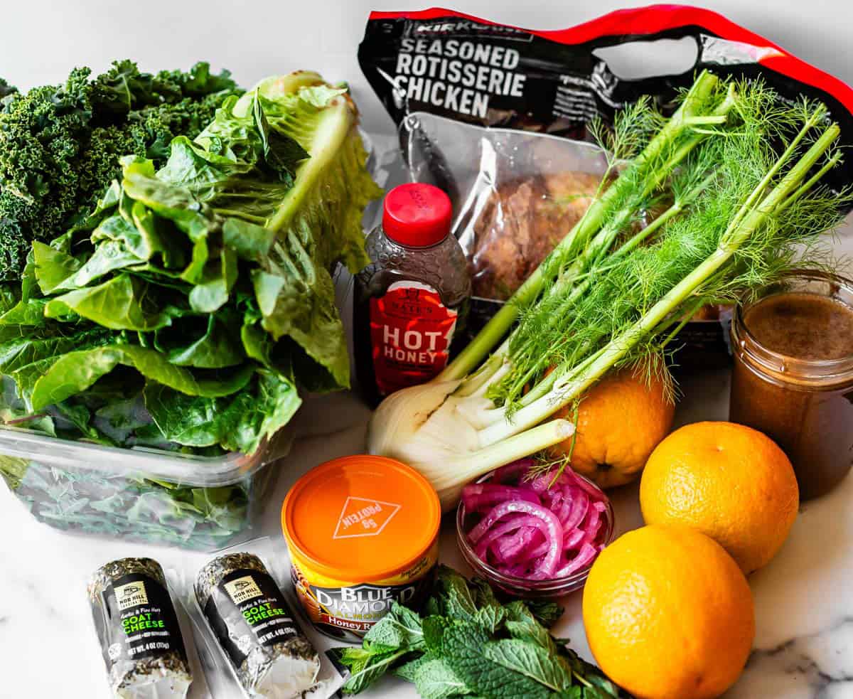 A variety of fresh ingredients including kale, romaine, fennel, oranges, mint, blue cheese, rotisserie chicken, pickled onions, hot honey, and almonds, arranged on a white surface.