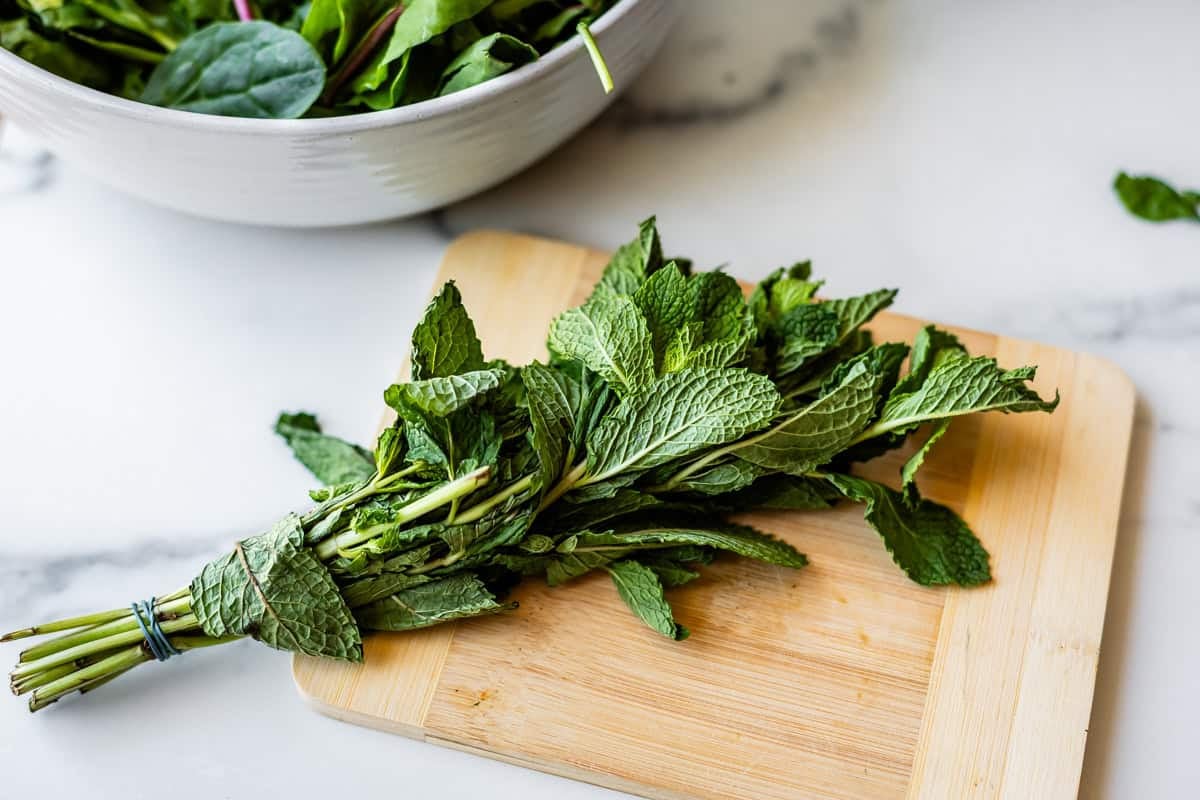 A bunch of fresh mint leaves tied with a rubber band is placed on a wooden cutting board, with a bowl of leafy greens in the background on a marble countertop.