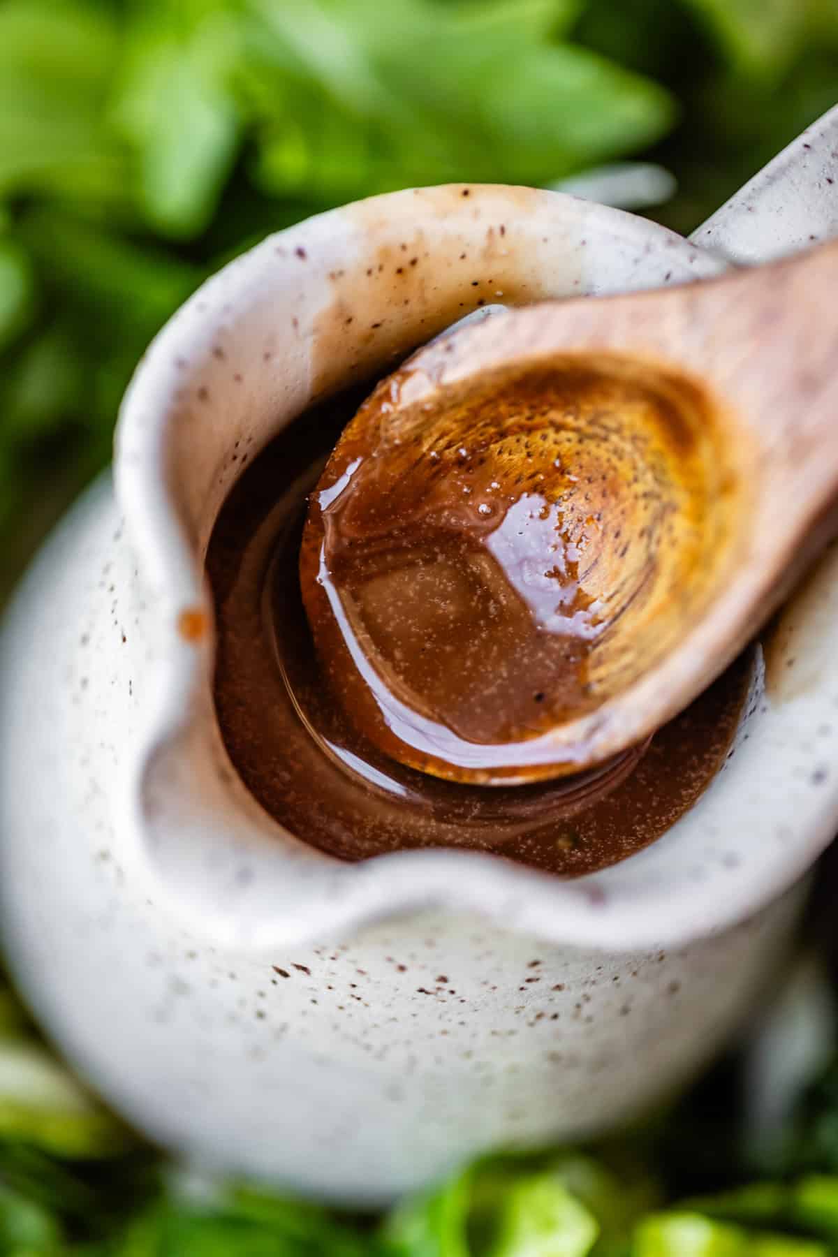 A close-up of a ceramic pitcher filled with brown vinaigrette dressing, with a wooden spoon resting inside. Fresh green leafy vegetables are blurred in the background.