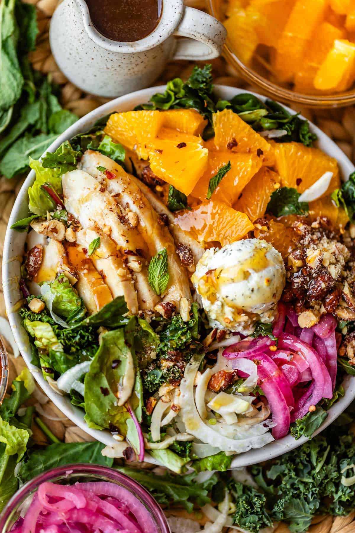 A colorful salad bowl with sliced grilled chicken, orange segments, mixed greens, pickled onions, cheese, nuts, and fresh herbs, served with a small jug of dressing and a bowl of orange slices nearby.