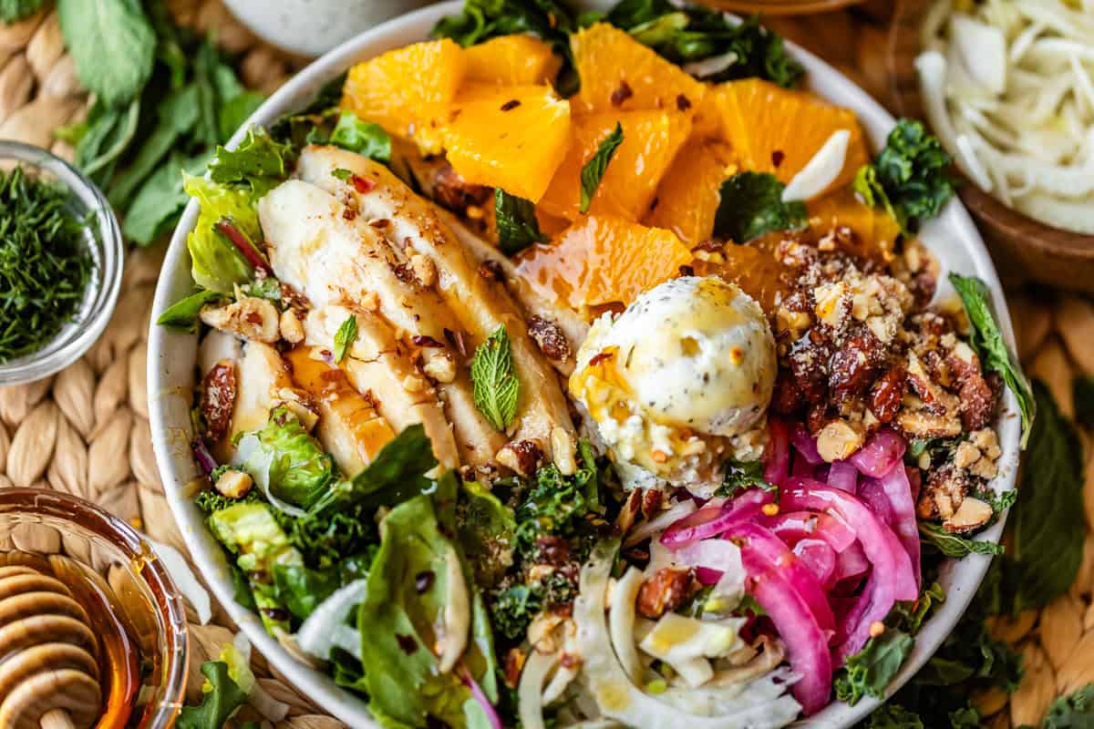 A colorful salad bowl with sliced chicken, orange segments, pickled onions, mixed greens, sliced fennel, nuts, and a dollop of creamy dressing, garnished with fresh herbs.
