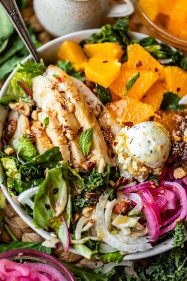 A salad bowl with grilled chicken, orange slices, pickled onions, greens, nuts, and a scoop of herbed cheese, garnished with fresh mint.