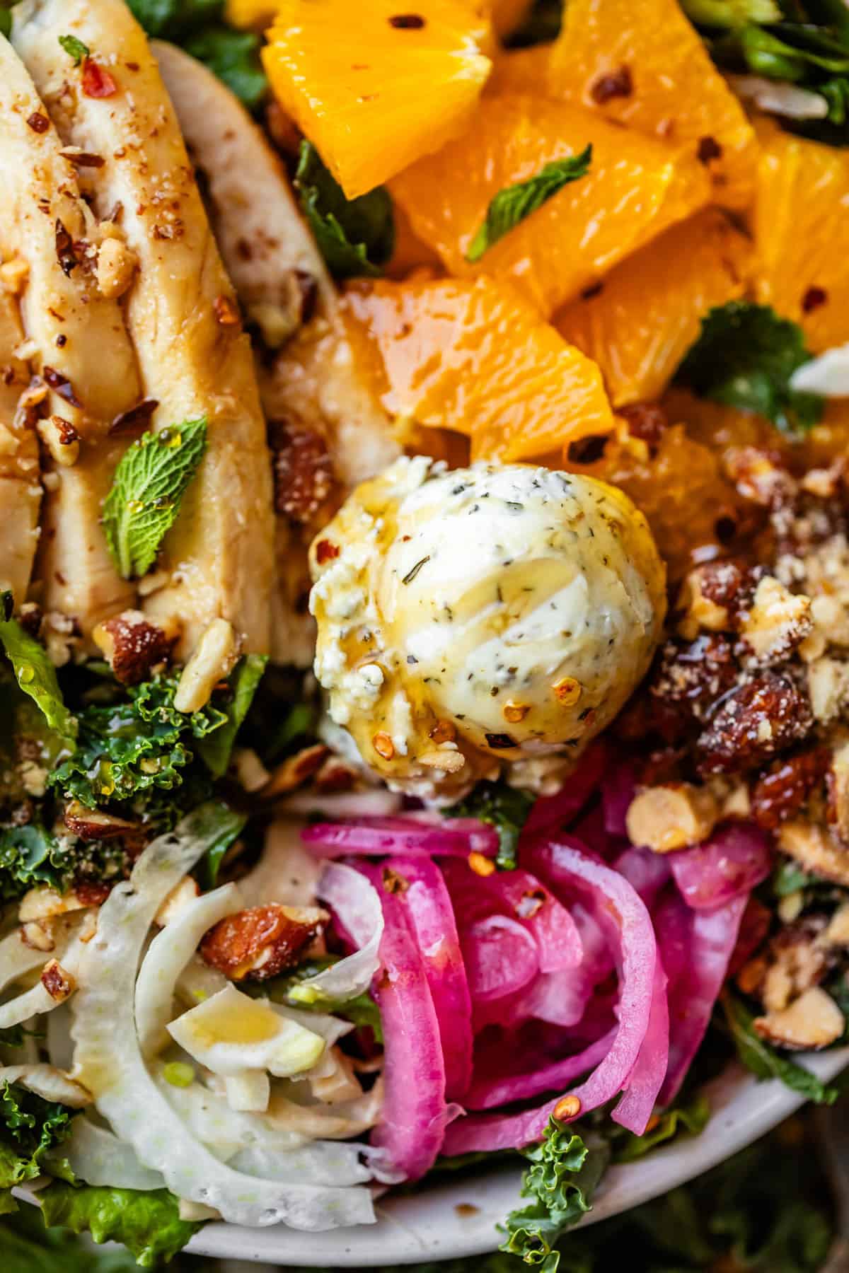 A close-up of a vibrant salad with sliced chicken, orange segments, goat cheese, pickled onions, fennel, kale, nuts, and fresh mint leaves.