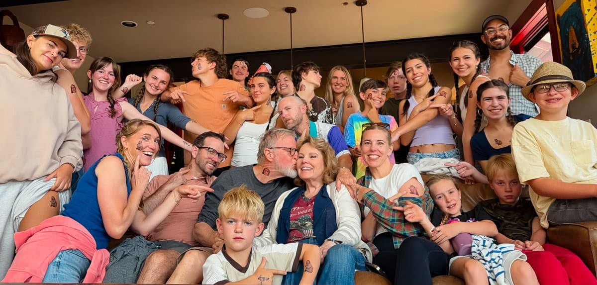 A large group of people, young and old, gather indoors, smiling and posing closely together, some showing off matching temporary tattoos on their arms, creating a lively and joyful atmosphere.