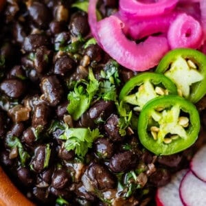 A bowl of black beans garnished with cilantro, sliced jalapeños, pickled red onions, and radish slices.